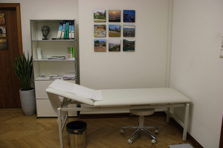 Eine Untersuchungsliege in einem Behandlungsraum mit einem Schrank und Bildern an der Wand.